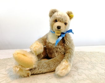 Pode incluir: Um urso de peluche vintage com pelo marrom claro, uma fita azul em volta do pescoço e um nariz de botão. O urso está sentado em um tecido de renda branca.