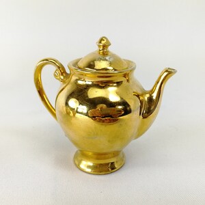 May include: A small, gold-colored teapot with a shiny, metallic finish. The teapot has a rounded body and a spout on one side. The lid is also gold-colored and has a knob on top.