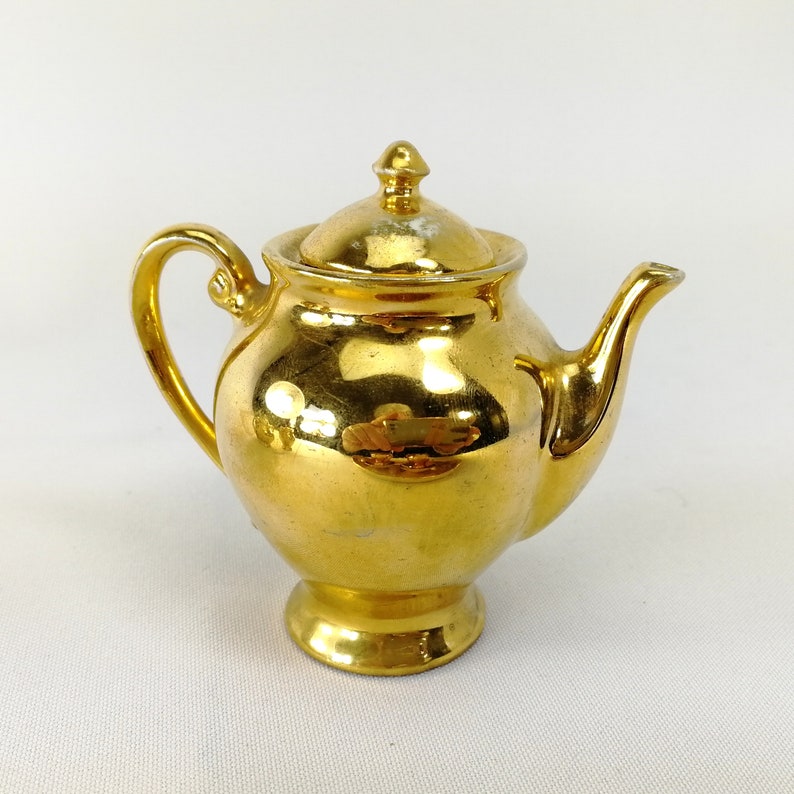 May include: A small, gold-colored teapot with a shiny, metallic finish. The teapot has a rounded body and a spout on one side. The lid is also gold-colored and has a knob on top.