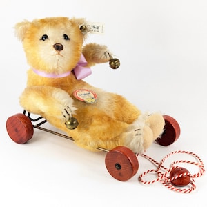 Puede incluir: Un oso de peluche Steiff vintage para tirar, con pelaje marrón claro y crema. El oso tiene una cinta rosa, campanas y ruedas de madera. Se adjunta un cordón rojo y blanco. El oso tiene una etiqueta Steiff.