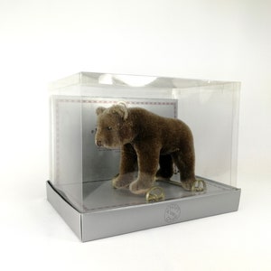 Puede incluir: Un oso de peluche marrón sobre ruedas en una vitrina de plástico transparente. El oso está sentado sobre una base blanca con un marco de metal dorado. La vitrina tiene una base plateada con el texto "KNOF HAMBURG" impreso en ella.