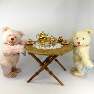 May include: Two teddy bears, one pink and one white, are having tea party with a gold tea set on a wooden table. The tea set includes a teapot, creamer, sugar bowl, and two teacups.