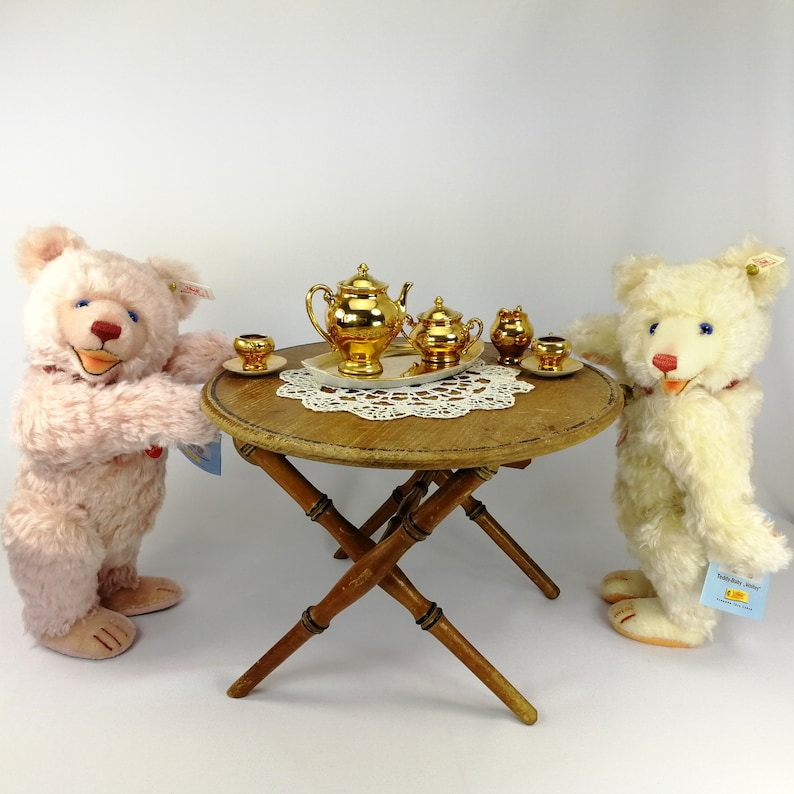 May include: Two teddy bears, one pink and one white, are having tea party with a gold tea set on a wooden table. The tea set includes a teapot, creamer, sugar bowl, and two teacups.