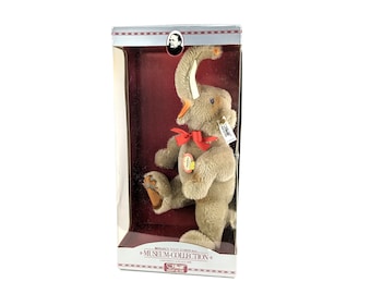 May include: A gray plush elephant toy with a red bow and a Steiff tag. The elephant is standing on its hind legs and has its trunk raised. The toy is in a clear plastic box with a red background. The box has the words "Steiff Museum Collection" printed on it.