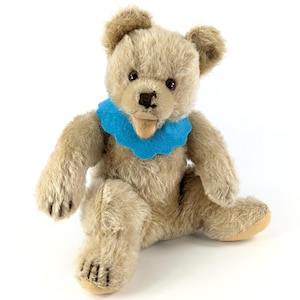 May include: A vintage teddy bear with a blue felt collar. The bear has a light brown fur and black button eyes.