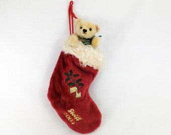 May include: Red plush Christmas stocking with a white fur cuff. A small teddy bear is peeking out of the top of the stocking. The stocking has an embroidered holly sprig and bells with the text "Steiff 2004" at the bottom.