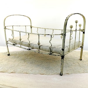 K&ouml;nnte beinhalten: Ein wei&szlig;es Puppenbett aus Metall mit einer wei&szlig;en Stoffmatratze. Das Bett hat ein gebogenes Kopfteil und Fu&szlig;teil mit dekorativen Metallkugeln.
