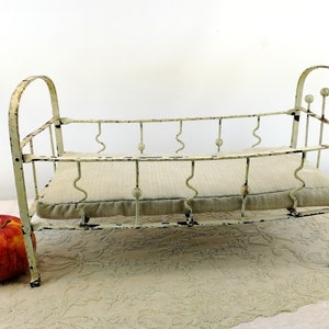 K&ouml;nnte beinhalten: Ein wei&szlig;es Puppenbett aus Metall mit einer wei&szlig;en Stoffmatratze. Das Bett hat ein gebogenes Kopfteil und Fu&szlig;teil mit dekorativen Metallkugeln.
