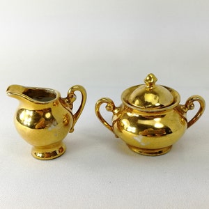 May include: A gold-plated porcelain creamer and sugar bowl set. The creamer has a handle and a spout. The sugar bowl has a lid and two handles.