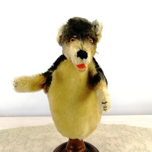 May include: A vintage hand puppet of a wolf, made of plush fabric. The wolf has a black and brown fur pattern, a black nose, and an orange mouth. The puppet is mounted on a wooden stand.