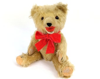 Könnte beinhalten: Ein Vintage-Teddybär mit einer roten Samt Schleife. Der Bär hat ein hellbraunes Fell und ein lächelndes Gesicht mit einem roten Mund.