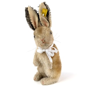 May include: A vintage plush rabbit toy with a tan body, long ears with dark tips, and a white ribbon bow. The rabbit has a small yellow tag attached to its ear. The toy is standing upright on a white surface.