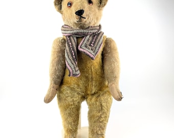 Pode incluir: Um urso de pelúcia vintage com corpo de pele castanho claro e nariz castanho escuro. O urso usa um cachecol com listras rosa, cinza e marrom. O urso está em um suporte branco sobre um fundo branco.