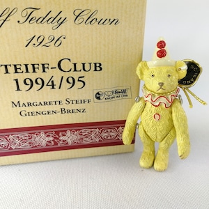 Könnte beinhalten: Ein gelber Teddybär, der als Clown verkleidet ist, mit einem rot-weißen Rüschenkragen, roten Bommeln auf seinem Hut und einem Steiff-Knopf im Ohr. Der Bär steht auf einer Schachtel, auf der "Steiff Teddy Clown 1926" und "Teiff-Club 1994/95" steht.