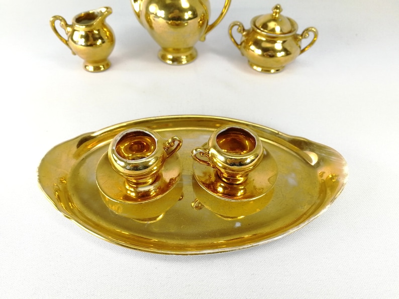 May include: A gold-colored tea set with a tray, teapot, creamer, sugar bowl, and two cups. The set is made of porcelain and has a shiny, metallic finish.