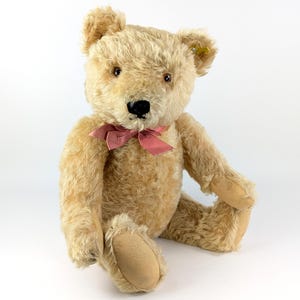 Op de afbeelding: Een vintage teddybeer met lichtbruine vacht, een zwarte neus en bruine ogen. De beer heeft een roze strik om zijn nek. De beer zit met uitgestrekte benen en armen langs zijn zij. De beer heeft bruinachtige poten.