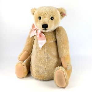 May include: A plush teddy bear with light brown fur, a black nose, and dark eyes. It has a pink satin bow tied around its neck and tan-colored paws. A small tag is attached to its ear.