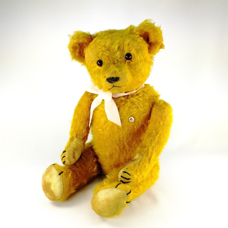Old yellow teddy bear sitting before a white background and smiling in the camera. He wears a white bow. His glass eyes are catching the light. Bear has a white identifying milk glass button on the left side of his chest.