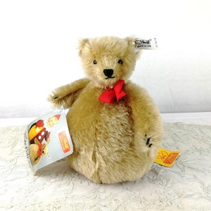 Puede incluir: Un oso de peluche Steiff marrón claro con un lazo rojo alrededor del cuello. El oso está sentado sobre un mantel de encaje blanco. El oso tiene una etiqueta que dice "Steiff" y "Hecho en Alemania".