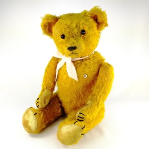 An old yellow teddy bear sitting before a white background and smiling in the camera. He wears a white bow. His glass eyes are catching the light. Bear has a white identifying milk glass button on the left side of his chest.