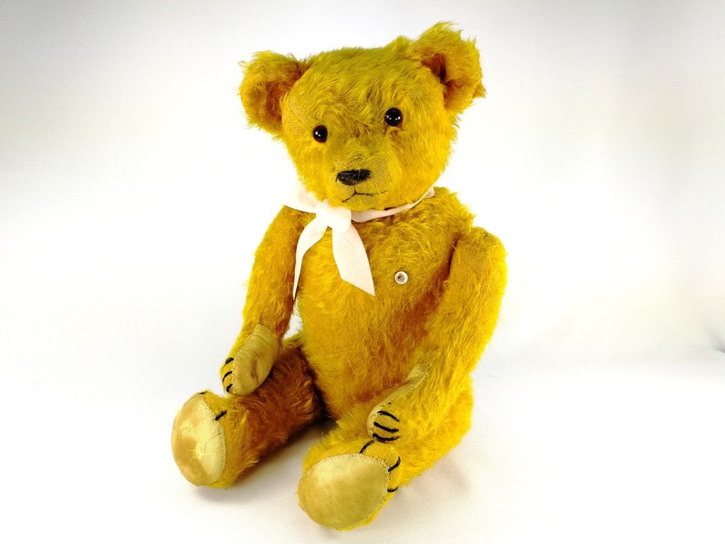 An old yellow teddy bear sitting before a white background and smiling in the camera. He wears a white bow. His glass eyes are catching the light. Bear has a white identifying milk glass button on the left side of his chest.