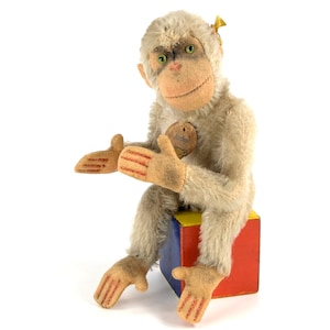 May include: A vintage plush monkey toy sitting on a colourful block. The monkey is beige with a brown face and green eyes. The monkey has a tag that says "Steiff" on its chest.
