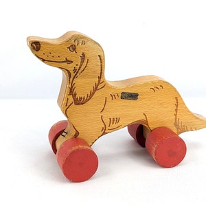 Könnte beinhalten: Hölzernes Spielzeug-Dackel mit roten Rädern. Der Dackel ist braun bemalt und hat eine schwarze Nase und Augen. Das Spielzeug ist aus Holz und hat ein einfaches Design.