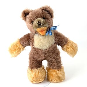 Puede incluir: Un oso de peluche marrón con un hocico marrón claro y un lazo azul. El oso tiene un pecho marrón claro y pelaje marrón claro en sus patas y brazos.