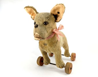 May include: A vintage plush toy French bulldog on wheels. The toy is gray with a pink bow and has wooden wheels.