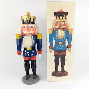 Op de afbeelding: Een houten notenkraker soldaat met een blauw en rood uniform, een witte baard en een gouden kroon. De notenkraker staat op een houten voet en houdt een notenkraker in zijn rechterhand. De notenkraker zit in zijn originele doos.