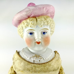 Può includere: Una bambola di porcellana con occhi azzurri, guance rosate e capelli biondi ricci. La bambola indossa un cappello rosa e un vestito beige.