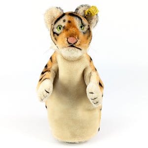 May include: A plush tiger hand puppet featuring orange and black stripes, a cream-colored chest, green eyes, and a pink stitched nose. A yellow Steiff brand tag is attached to the left ear with a metal button, displaying the text 0860.17.