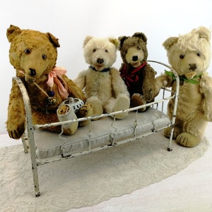 K&ouml;nnte beinhalten: Vier vintage Teddyb&auml;ren sitzen auf einem wei&szlig;en Metallbettgestell mit einer wei&szlig;en Matratze. Die B&auml;ren sind alle in verschiedenen Farben und haben unterschiedliche Gesichtsausdr&uuml;cke.