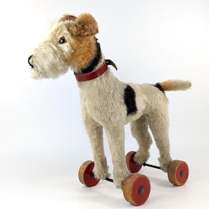 Puede incluir: Un perro de juguete vintage con cuerpo color crema, cabeza marrón y beige, y marcas negras. El perro tiene un collar rojo y está montado sobre cuatro ruedas de madera con centros rojos. Un juguete clásico para niños.