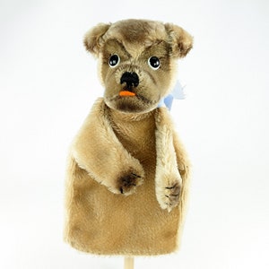 May include: A brown plush hand puppet shaped like a dog with a blue ribbon around its neck. The puppet has black eyes, a black nose, and an orange mouth.