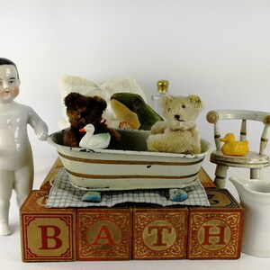 May include: A white porcelain doll with black hair stands next to a toy bathtub with a brown teddy bear, a green frog, and a white duck inside. The bathtub is on top of wooden blocks that spell out "BATH".
