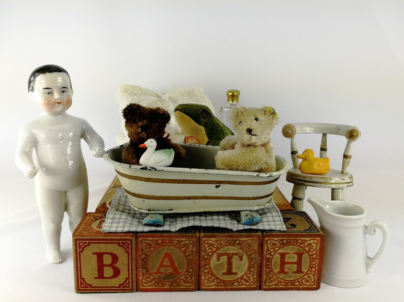 May include: A white porcelain doll with black hair stands next to a toy bathtub with a brown teddy bear, a green frog, and a white duck inside. The bathtub is on top of wooden blocks that spell out "BATH".