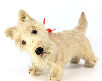 May include: A plush toy dog with cream-colored fur, pointed ears, and dark brown eyes. The dog has a black stitched nose and mouth, and a small red ribbon tied around its neck. It is standing in a slightly tilted position with its tail raised.