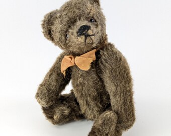 May include: A vintage brown teddy bear with a tan bow tie. The bear has a classic design with a stitched nose and mouth. The fur is a dark brown color, and the bear is sitting with its arms and legs slightly bent.