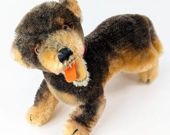 Könnte beinhalten: Ein braunes und braunes Plüschspielzeug-Hund mit einem roten Halsband und einer orangefarbenen Zunge, die herausragt. Der Hund liegt auf dem Bauch mit dem Kopf zur Seite gedreht.