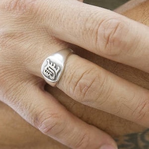 May include: A silver signet ring with a raised design on the top. The ring is worn on the index finger of a person's hand.