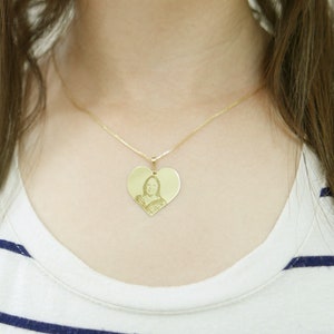 May include: A gold heart-shaped pendant necklace with a photo of a person inside. The necklace is worn by a person with long brown hair.