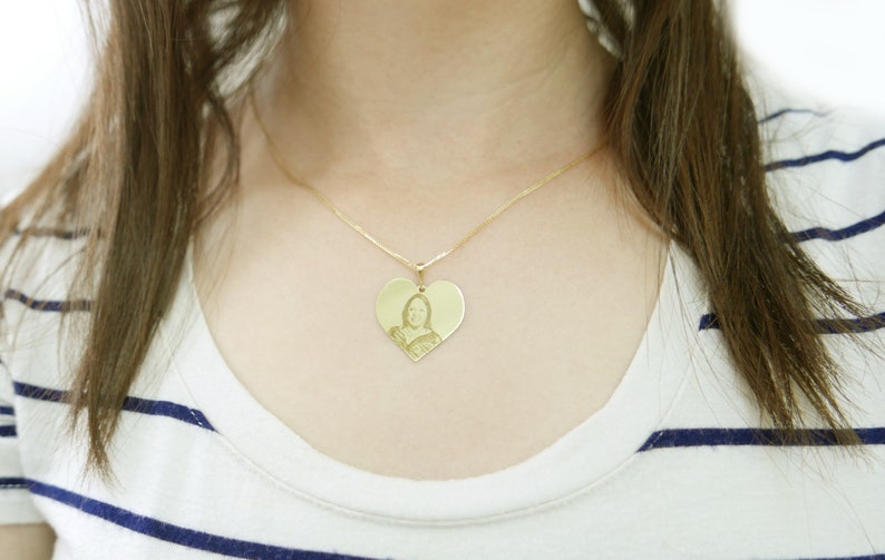 May include: A gold heart-shaped pendant necklace with a photo of a person inside. The necklace is worn by a person with long brown hair.