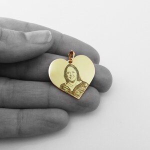 May include: Gold heart-shaped pendant with an engraved image of a person. The pendant is in the palm of a hand.