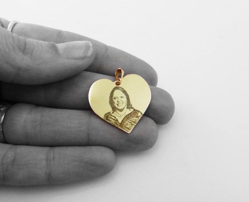 May include: Gold heart-shaped pendant with an engraved image of a person. The pendant is in the palm of a hand.
