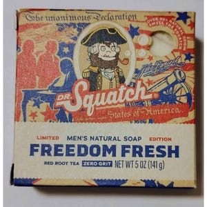 May include: A rectangular box of Dr. Squatch Freedom Fresh natural soap. The packaging features a patriotic theme with red, white, and blue colors, a cartoon character in a tricorn hat, and text including 'Freedom Fresh', 'Red Root Tea', and 'Net Wt 5 oz'.