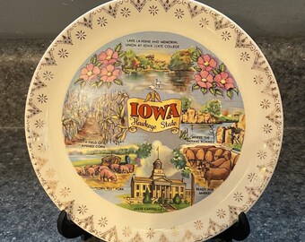May include: A white ceramic plate with a gold border and a colorful illustration of Iowa. The plate features images of cornfields, a state capitol building, and a lake. The text "Iowa Hawkeye State" is printed in the center of the plate.