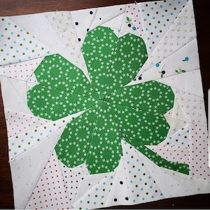 May include: A green four-leaf clover fabric block with a white background. The clover is made of green fabric with a white star pattern. The background is white with a variety of small, colorful polka dots.