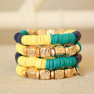 Op de afbeelding: Een stapel van vier armbanden met gele, turquoise en donkerblauwe kralen. De armbanden zijn versierd met gouden letterkralen die het woord "UNC" twee keer spellen.