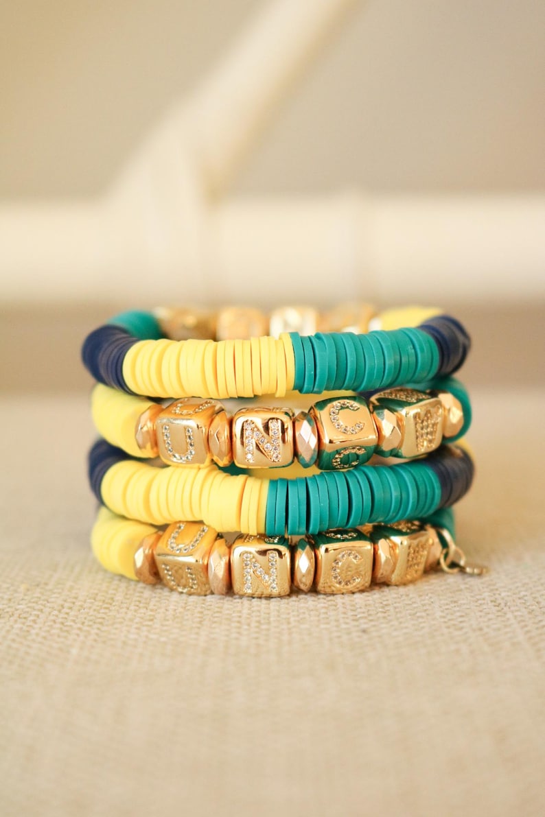 Op de afbeelding: Een stapel van vier armbanden met gele, turquoise en donkerblauwe kralen. De armbanden zijn versierd met gouden letterkralen die het woord "UNC" twee keer spellen.